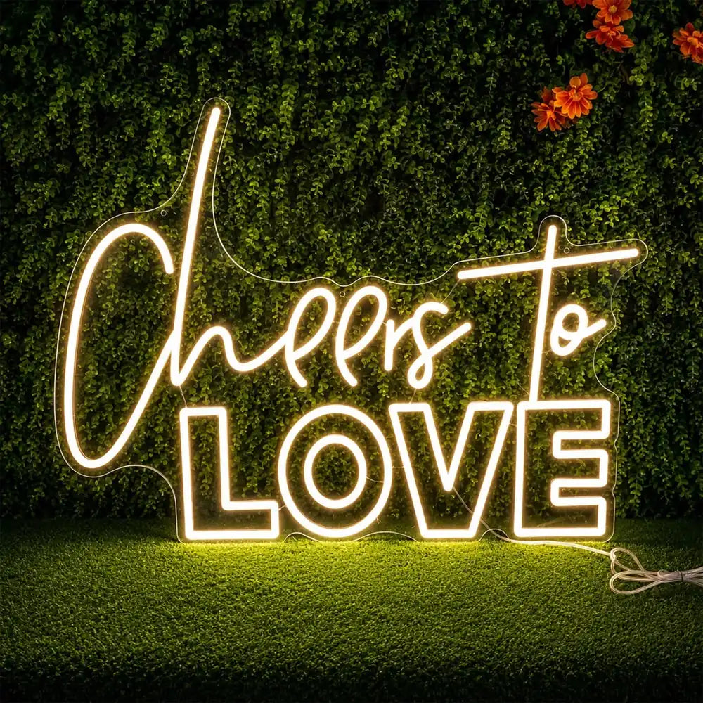 Warmweißes Neonschild mit dem Schriftzug Cheers to Love vor einer Pflanzenwand